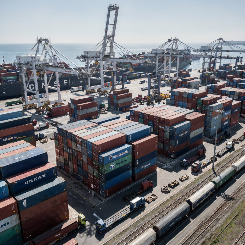 An aerial view of a busy container port showing stacked containers, gantry cranes operating, trucks and trains moving, clear skies, no text
