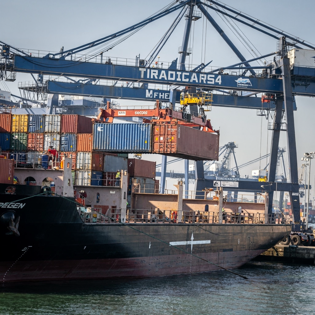 Close-up view of a dual-lane quay crane lifting a container from a vessel, with nearby yard cranes and trucks visible, shot in clear conditions without text or numbers