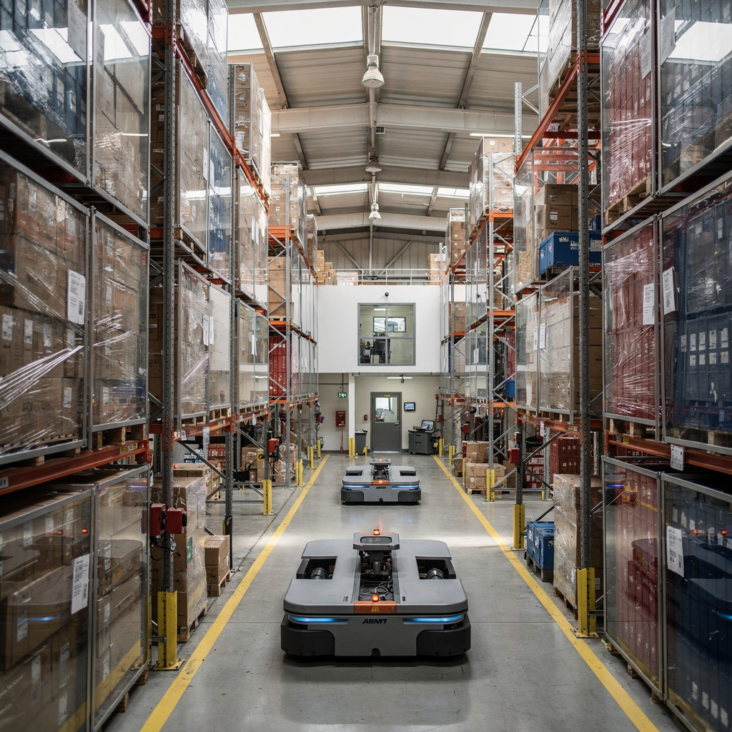 Retrofitted warehouse yard with AGVs and adapted aisles Interior view of a retrofitted warehouse yard showing AGVs moving containers, narrowed aisles adapted for autonomous vehicles, storage racks and a small control room, bright lighting, no text