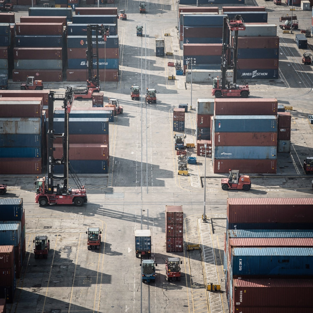An aerial view of a container terminal storage yard showing rows of stacked containers, automated guided vehicles and yard cranes working, sunny day, no text